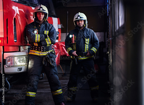 Two firemen wearing protective uniform standing next to a fire engine in a garage of a fire department.