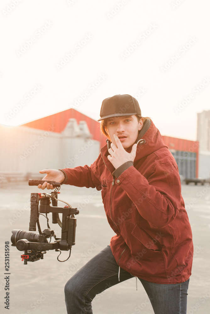 Funny man with a stabilizer and camera on the background of the sunset. Portrait of a young videographer holding a stabilizer in his hands and posing on the camera.
