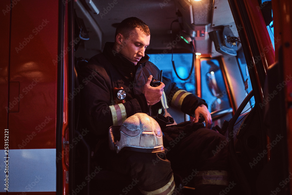 The fire brigade arrived at the night-time. Fireman in a protective uniform sitting in the fire truck and talking on the radio The fire brigade arrived at the night-time. Fireman in a protective uniform sitting in the fire truck and talking on the radio