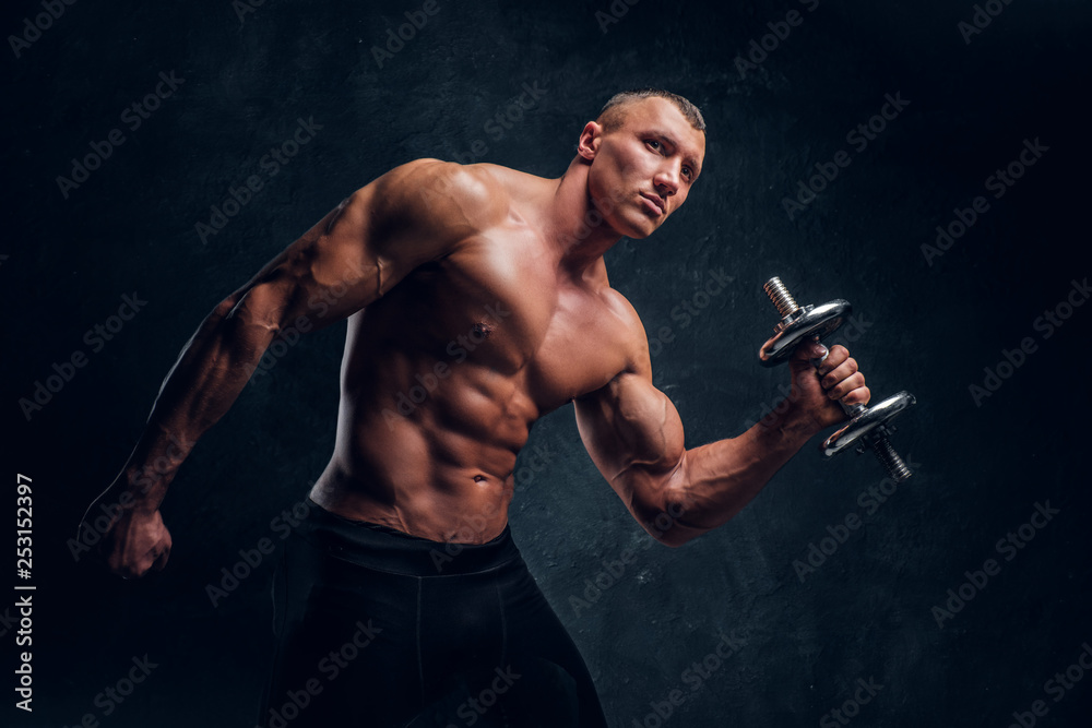 A young guy with a perfect pumped body posing with a dumbbell. Studio photo with dark wall background