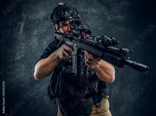 Bearded special forces soldier wearing body armor and helmet with night vision holding an assault rifle. Studio photo against a dark textured wall