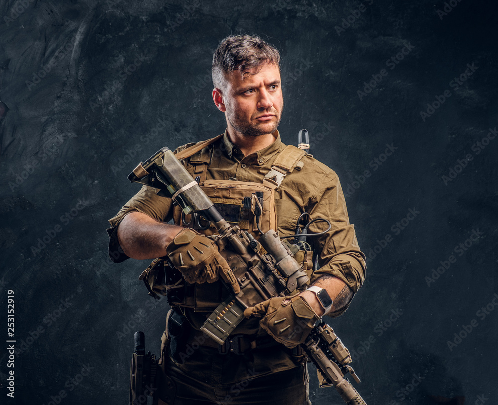 Foto Stock Special forces soldier wearing body armor holding assault rifle.  Studio photo against a dark textured wall | Adobe Stock