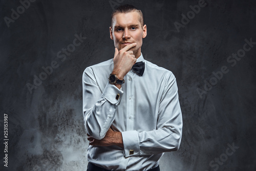 Confident elegantly dressed man wearing white shirt and bow tie, touching his face and looking at a camera. Studio shot on a dark textured wall