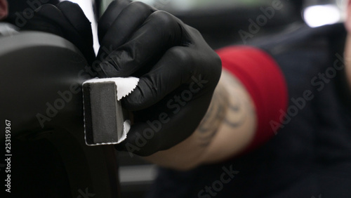A professional worker guy (Male) applies ceramics (special liquid) to the car using an applicator (sponge) in black gloves and in a protective robe. Concept of: Auto service, Deteyling.