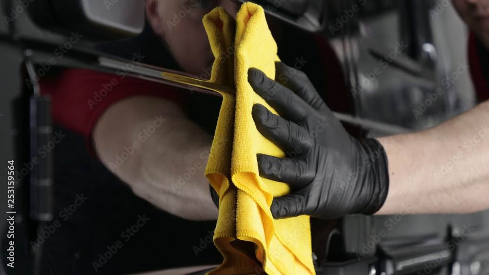 Professional worker guy (Man) wipes the car after painting (ceramics) in a special uniform with black gloves and with a yellow rag. Concept of: Young master, Preparing for the sale, Cleaning.