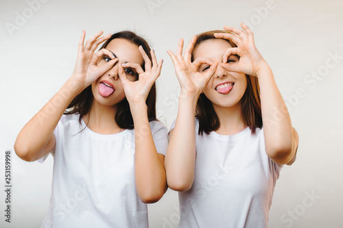 Portrait of two pretty girls having fun and showing their tongues while looking thru fingers dressed in white shirts against white wall. photo