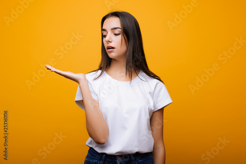 Beautiful young caucasian brunette looking at her hand amazed dressed in casual clothes in front of a yellow studio wall.