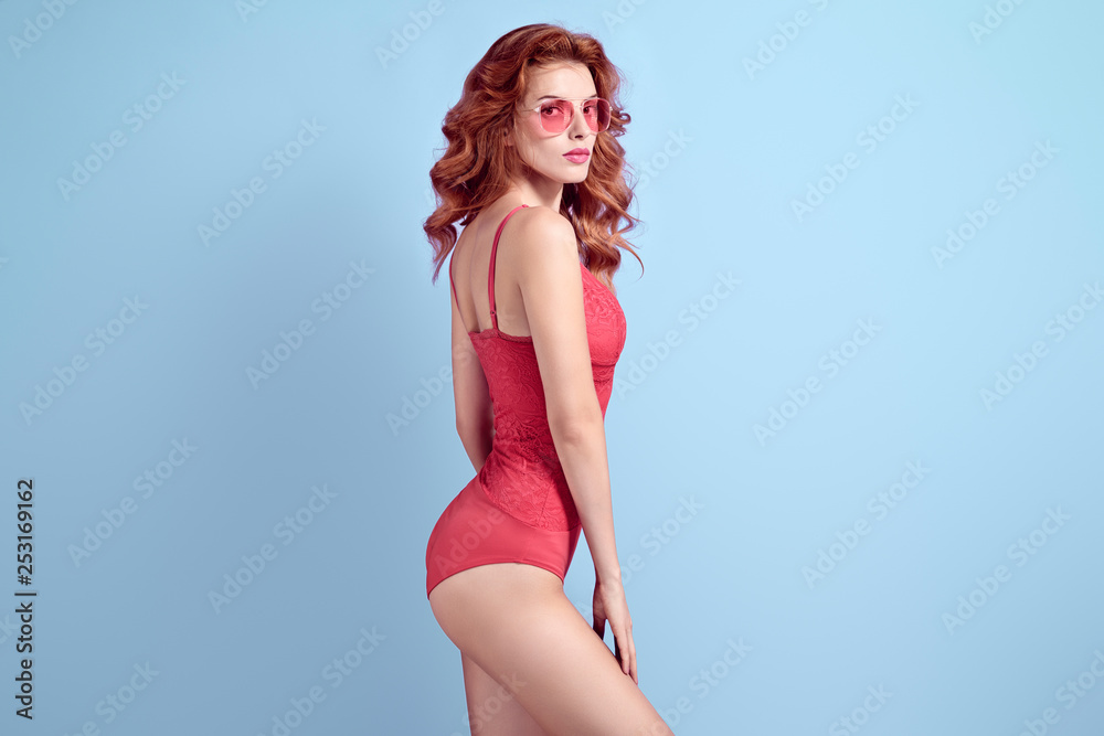 Fashionable shapely Redhead woman with make up wearing Stylish Coral  bodysuit. Beautiful sexy fitness Lady in Trendy lace Attire, fashion  Sunglasses. Slim Female model in Studio Stock Photo