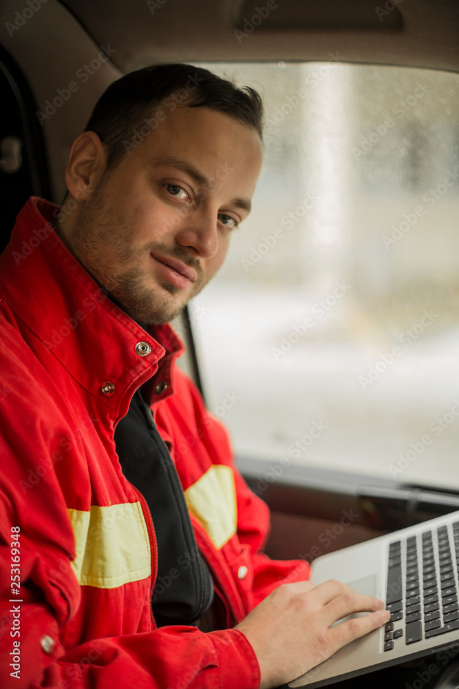 Young handsome paramedic in his ambulance smiling and writing report on his laptop. Looking at camera.
