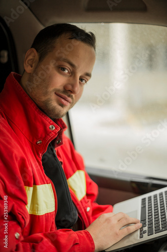 Obraz na płótnie Young handsome paramedic in his ambulance smiling and writing report on his laptop. Looking at camera. w salonie