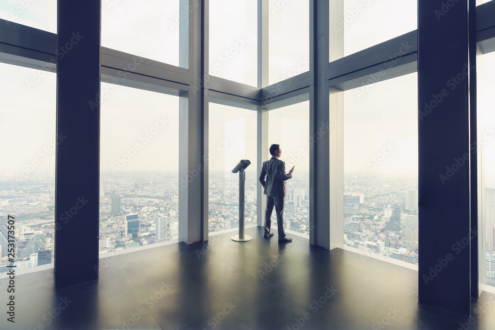 Business man stand by glass window in tall building with background of city Business man stand by glass window in tall building with background of city