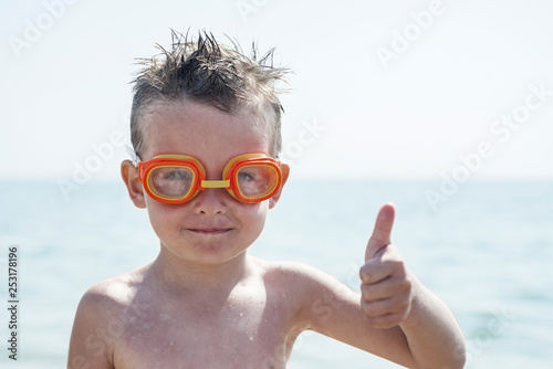 A boy in glasses for swimming shows a thumbs up on the background of the sea. Kid in spectacles for swimming in the sea © spyrakot