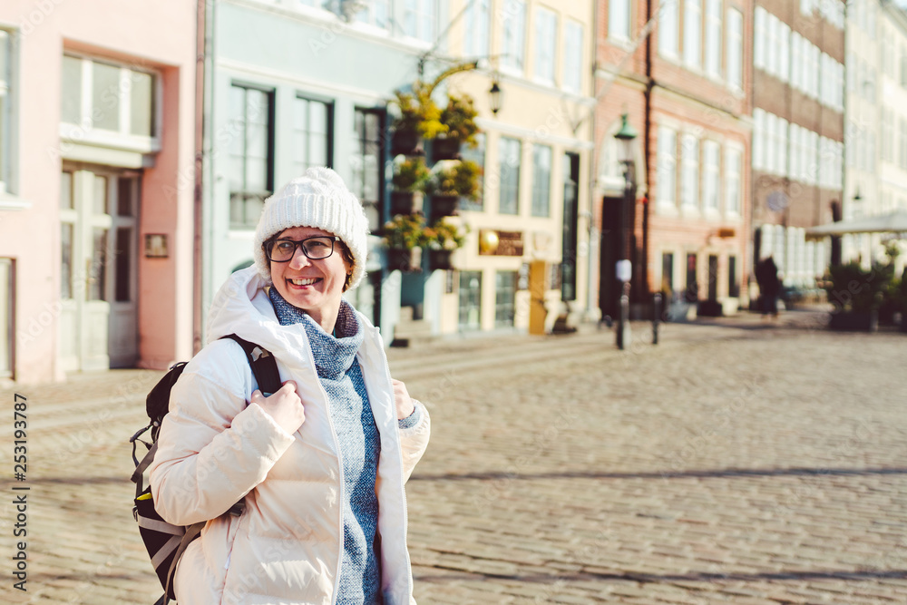 Portrait of a young Caucasian European woman tourist in glasses for the view of a white hat and a down jacket with a black backpack posing on an old Europe street in the city of Copenhagen in Denmark