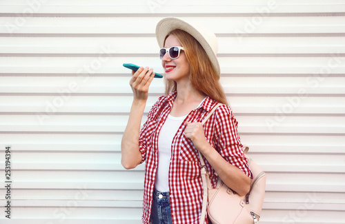 Portrait happy smiling woman holding phone using voice command recorder or calling, wearing summer round straw hat with bag walking on city street over white wall background