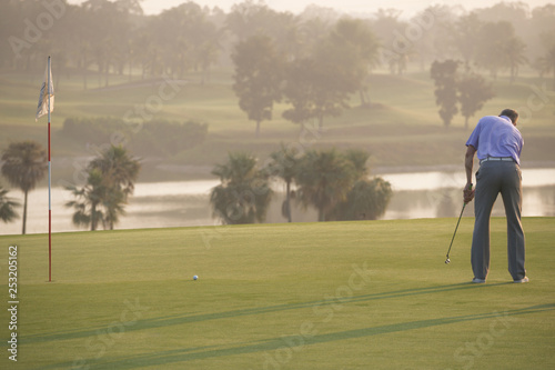 Golfers who are putting golf balls on the green in the warm sunshine in the morning. The view from the back. 