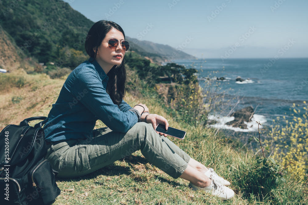 young girl backpacker travel trip outdoor adventure in big sur coastline california usa. beautiful lady in sunglasses holding mobile phone sitting on meadow relaxing sightseeing blue ocean view  sky