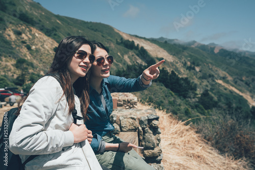 two active teenage female hikers on top of the mountain relaxing resting sitting on cliff sightseeing pointing view in big sur california usa. young girls travelers chatting talking cheerfully.