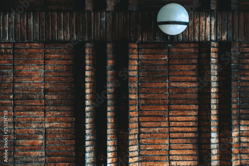 A dark atmospheric wall of oblong bricks near the house entrance with several triangle displacements; the dark texture of a brick wall with matte glass lamp inside of the porch of a residential house