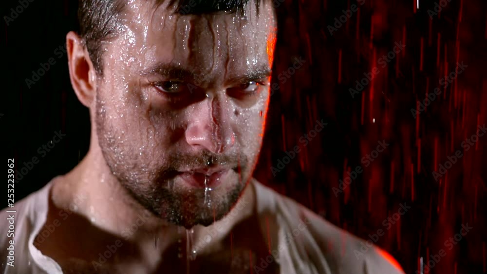 close up. portrait of a man in the rain in the dark. on the face of flowing jets and drops of water. angry look resentment red neon lighting