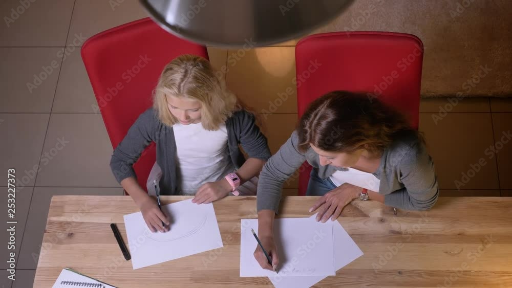 Closeup top view of small pretty blonde girl learning how to draw while mother giving her a lesson Closeup top view of small pretty blonde girl learning how to draw while mother giving her a lesson