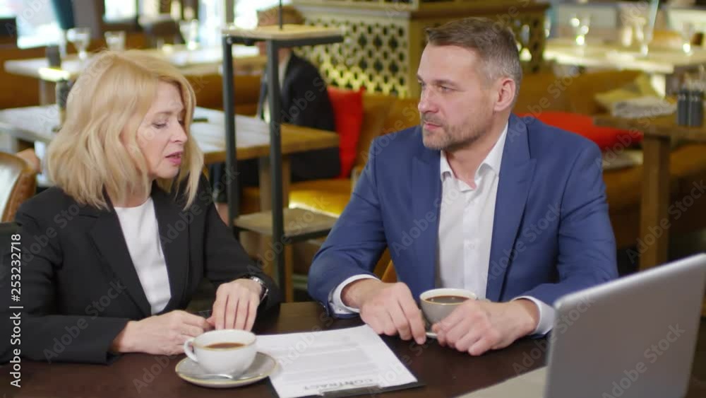 Medium shot of businessman and businesswoman sitting in restaurant, discussing contract lying on table and then shaking hands Medium shot of businessman and businesswoman sitting in restaurant, discussing contract lying on table and then shaking hands