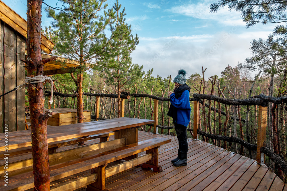 Front view of a girl at the top of a treetop wooden cabin glamping drinking some tea in the exterior with a beautiful view of the forest from the heights. Front view of a girl at the top of a treetop wooden cabin glamping drinking some tea in the exterior with a beautiful view of the forest from the heights.
