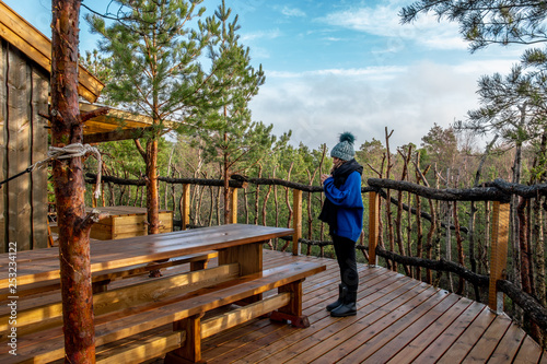 Front view of a girl at the top of a treetop wooden cabin glamping drinking some tea in the exterior with a beautiful view of the forest from the heights.