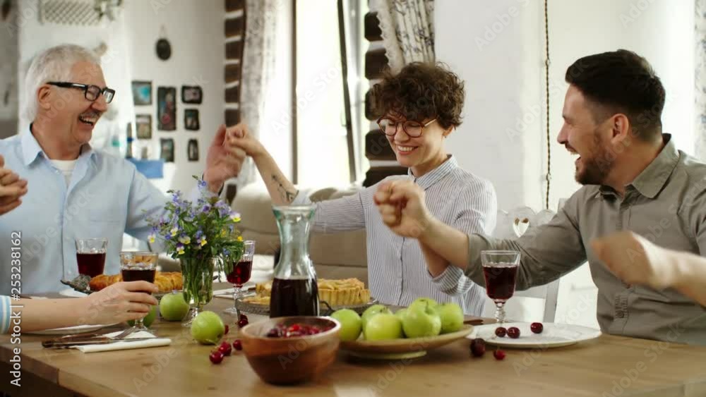 Medium shot of Caucasian family sitting at dinner table, holding hands, shaking them and singing joyfully Medium shot of Caucasian family sitting at dinner table, holding hands, shaking them and singing joyfully