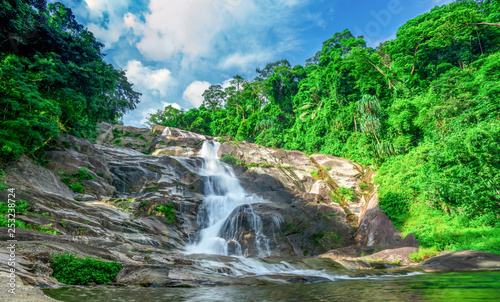 Beautiful waterfall at the mountain with blue sky and white cumulus clouds. Waterfall in tropical green tree forest. Waterfall is flowing in jungle. Nature abstract background. Granite rock mountain.