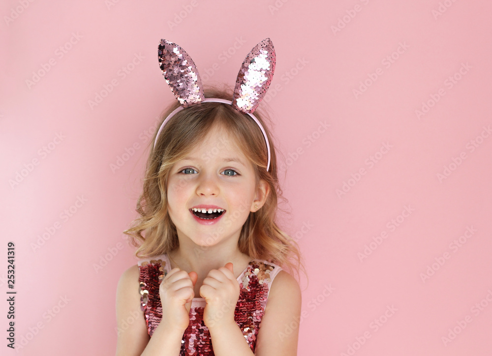 Cute little child wearing bunny ears on Easter day on pink background. Easter girl portrait, funny emotions, surprise. Copyspace for text.