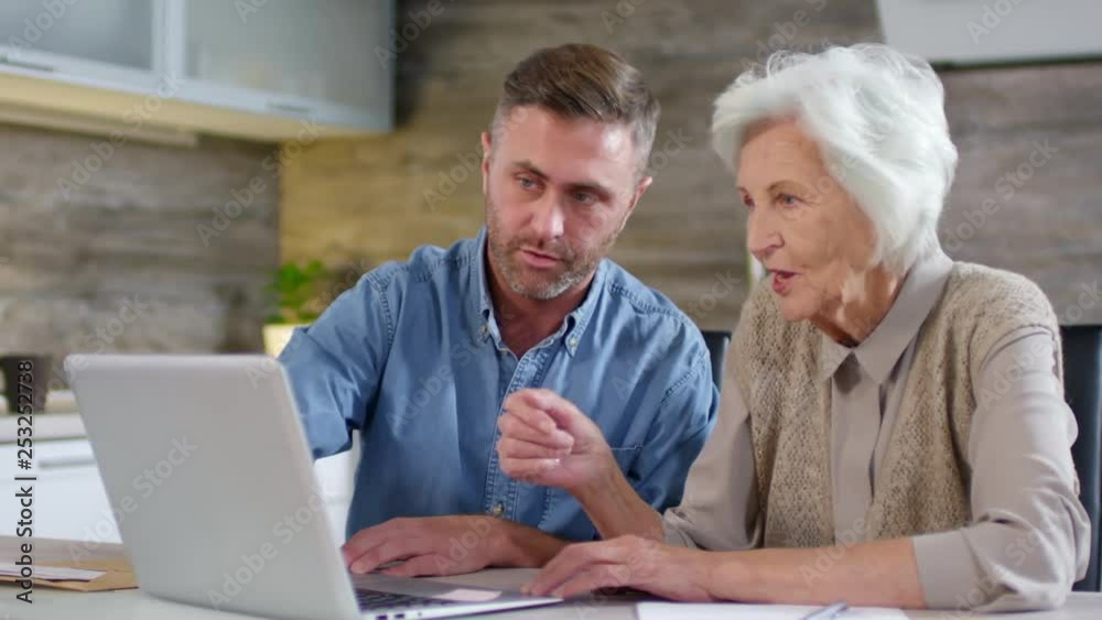 Lockdown of adult man sitting together with his aged mother at kitchen table and teaching her how to use computer Lockdown of adult man sitting together with his aged mother at kitchen table and teaching her how to use computer
