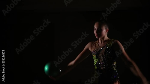 Slim cute attractive girl athlete in bright colorful swimsuit performs elements of rhythmic gymnastics with ball in dark gym with beautiful staged light and rear light