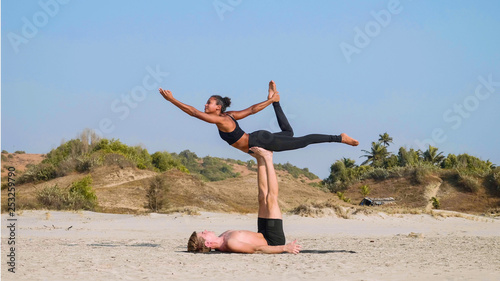 Fit sporty couple practicing acro yoga with partner together on the sandy beach. Mix race couple doing acrobatic exercise. Female acrobat is balancing on the legs of her male partner.