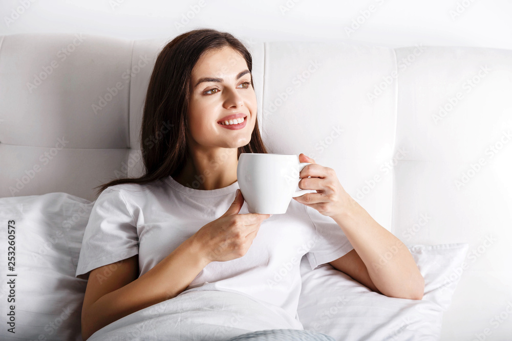 Happy young woman with cup of coffee or tea in bed at home bedroom