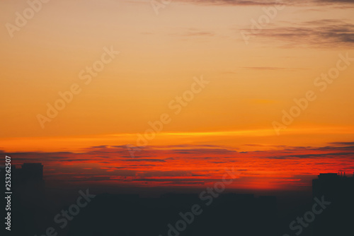 Cityscape with vivid fiery dawn. Amazing warm dramatic cloudy sky above dark silhouettes of city building roofs. Orange red sunlight. Atmospheric background of sunrise in overcast weather. Copy space.