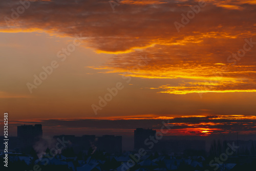 Cityscape with vivid fiery dawn. Amazing warm dramatic cloudy sky above dark silhouettes of city building roofs. Orange sunlight. Atmospheric background of sunrise in overcast weather. Copy space.