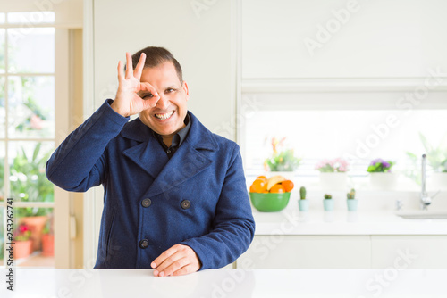 Middle age man sitting at home doing ok gesture with hand smiling, eye looking through fingers with happy face.