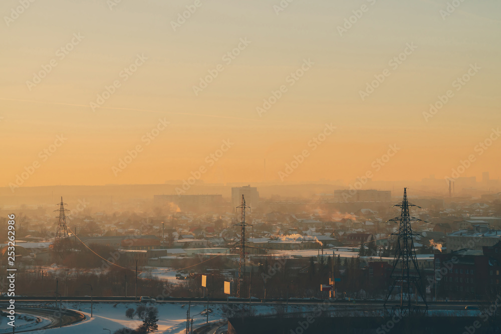 Power lines in city on dawn. Silhouettes of urban buildings among smog on sunrise. Cables of high voltage on warm orange yellow sky. Power industry at sunset. City power supply. Mist urban background.