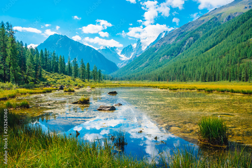Beautiful glacier reflected in mountain pure water with plants on bottom. Wonderful lake with snowy rocks reflection. White clouds on snowy mountains under blue sky. Amazing summer highland landscape.