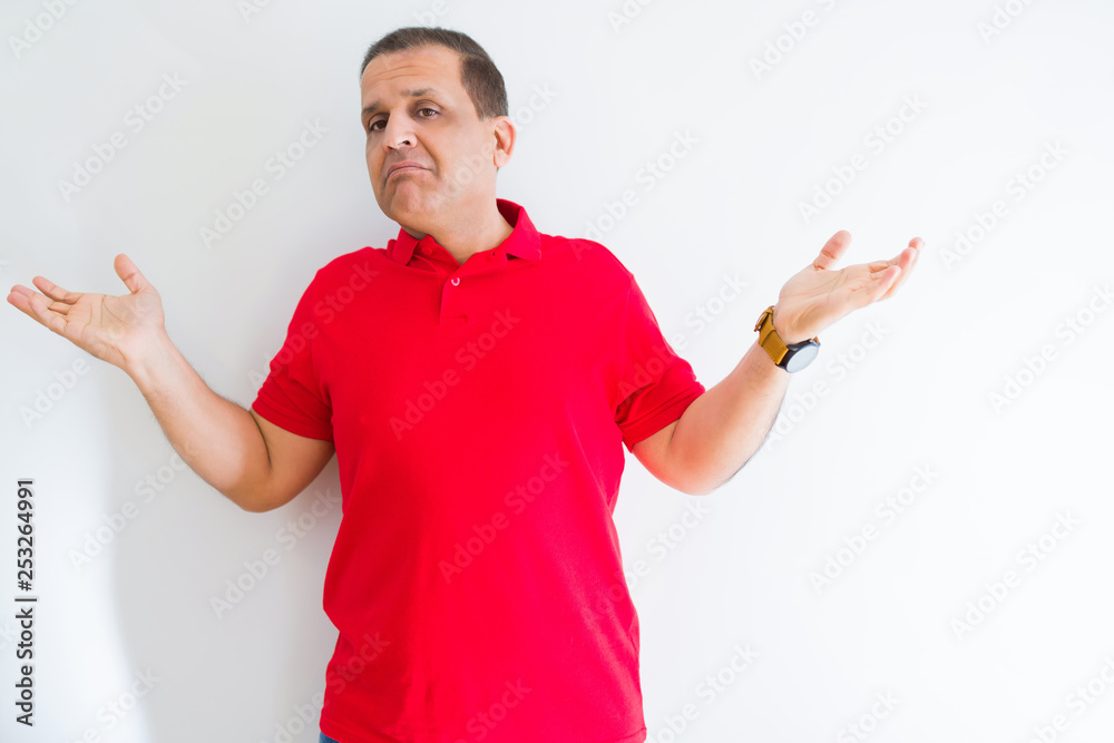 Middle age man wearing red t-shirt over white wall clueless and confused expression with arms and hands raised. Doubt concept. Middle age man wearing red t-shirt over white wall clueless and confused expression with arms and hands raised. Doubt concept.