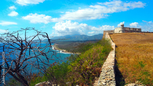 Wall of Castle San Pedro de la Roca del Morro, splitting the sea with a tree in front and the fortress in Santiago de Cuba, Cuba, February 2019
