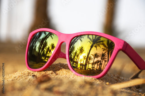 Pink modern sunglasses on the beach sand with palm trees and sunset reflection 