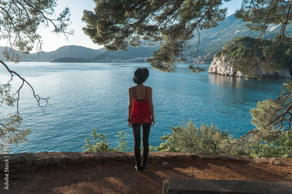 A girl stands under an old tree on a cliff with a beautiful view of the sea and mountains. Vacation in the European resort. Travel and leisure.