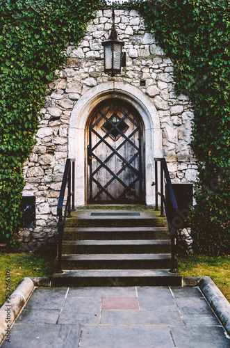 Antique arched wrought-iron doors in an old stone fortress wall using green lush ivy. Beautiful antique frnar over the old door