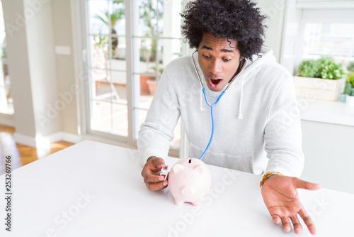 African American man holding stethoscope checking financial health on piggy bank scared in shock with a surprise face, afraid and excited with fear expression