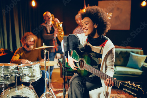 Mixed race woman singing and playing guitar while sitting on chair with legs crossed. In background drummer, saxophonist and bass guitarist. photo