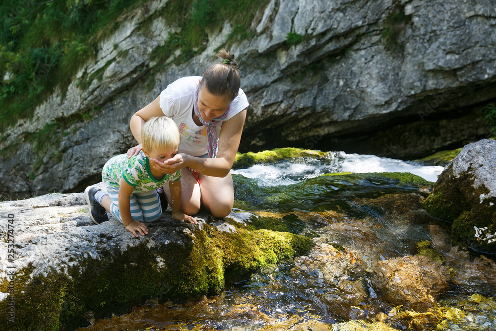 Mother with son drinking water from a pure, fresh and cool mountain stream on a family trip.