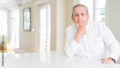 Handsome senior man at home looking confident at the camera with smile with crossed arms and hand raised on chin. Thinking positive.