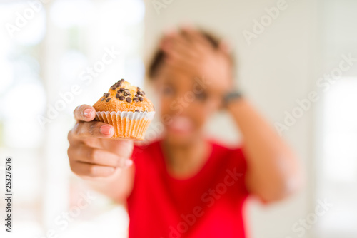 Young african american woman eating chocolate chips muffins stressed with hand on head  shocked with shame and surprise face  angry and frustrated. Fear and upset for mistake.