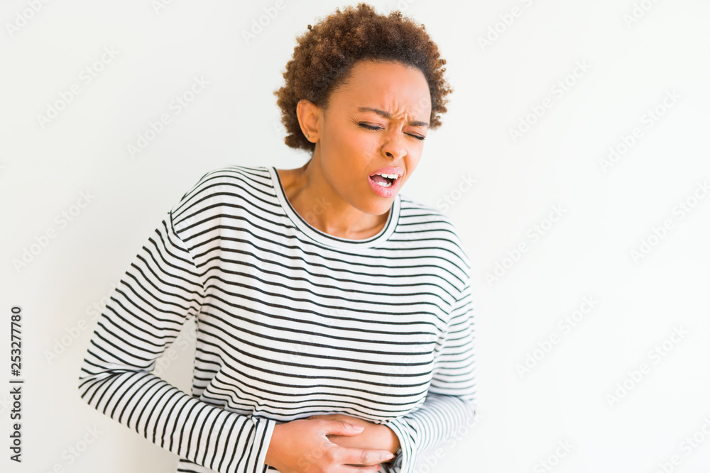 Young beautiful african american woman wearing stripes sweater over white background with hand on stomach because nausea, painful disease feeling unwell. Ache concept. Young beautiful african american woman wearing stripes sweater over white background with hand on stomach because nausea, painful disease feeling unwell. Ache concept.
